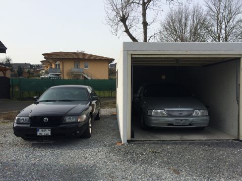 1999 Ford Fairlane - A black sedan is parked on a gravel driveway in front of a single-car garage. A silver sedan is parked inside the garage.