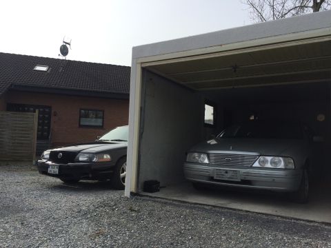 1999 Ford Fairlane - A black sedan is parked on a gravel driveway in front of a brick house and a garage. Another silver sedan is parked inside the open garage.
