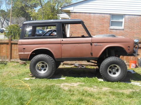 1972 Ford Bronco - A lifted 1972 Ford Bronco in a rusty brown color is parked on a grassy lawn in front of a brick house. The vehicle has large off-road tires…