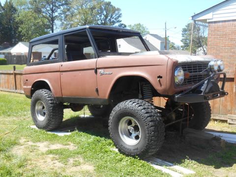 1972 Ford Bronco - A weathered, rusty brown 1972 Ford Bronco with a black hardtop is parked on a grassy lawn next to a brick building. The off-road vehicle fe…