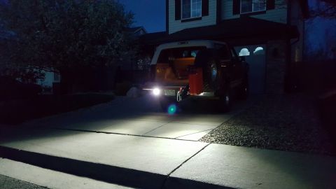 1996 Ford Bronco - The rear of a dark colored SUV is parked in a driveway at night, illuminated by its own bright reverse lights. A spare tire and a red gas c…