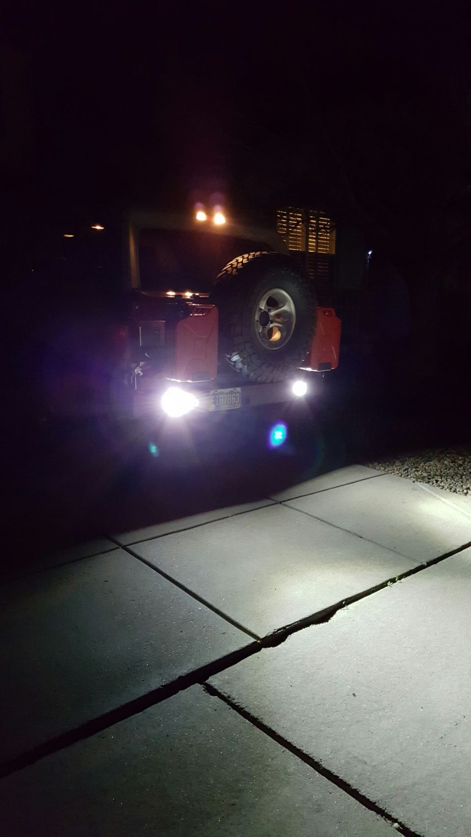 Rear view of a red SUV at night with bright LED lights, spare tire, and jerry cans.