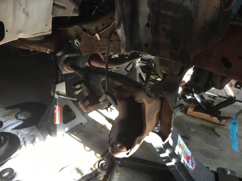 1987 Ford Bronco - The underside of a vehicle is shown, with the suspension components disassembled and supported by jack stands and a hydraulic jack. The sce…
