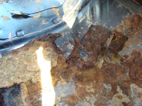 1981 Ford F-100 - The image shows severe rust and damage to the interior floor pan of a vehicle, with some carpet padding visible. A black cable and a small…