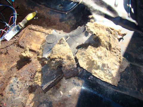 1981 Ford F-100 - The interior floor of a vehicle shows significant rust and damage, with torn insulation and exposed wiring. A scraper tool with a yellow ha…