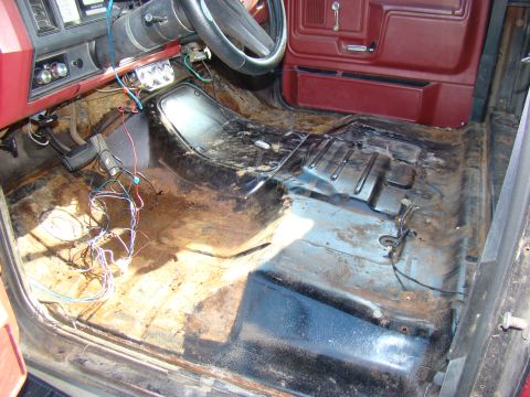 1981 Ford F-100 - The interior of a classic vehicle shows a stripped-down floor with visible rust and wiring. The dashboard, steering wheel, and a portion of…