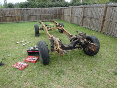 1981 Ford F-100 - A rusty 1981 Ford F-100 chassis sits on a grassy lawn in front of a wooden fence. Tools and shock absorbers are laid out on the grass nearb…