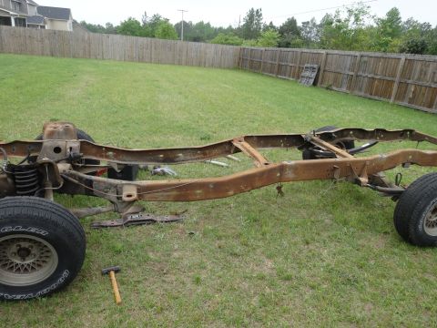 1981 Ford F-100 - The rusty frame of a pickup truck sits on a grassy lawn in front of a wooden fence. The front tire and suspension components are visible on…