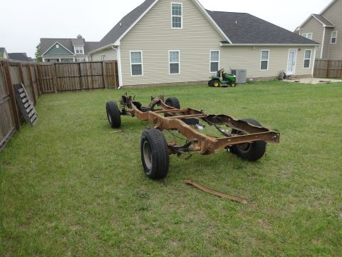 1981 Ford F-100 - The rusty frame of a pickup truck sits on a grassy lawn in front of a house. The truck frame has four wheels and suspension components visi…