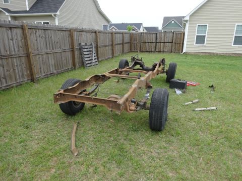 1981 Ford F-100 - A rusty truck frame with wheels and suspension components sits on a grassy lawn in front of a wooden fence and houses. Two shock absorbers…