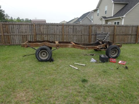 1981 Ford F-100 - A rusty, disassembled truck frame with wheels sits on a grassy lawn in front of a wooden fence and houses. Tools and parts are scattered ar…