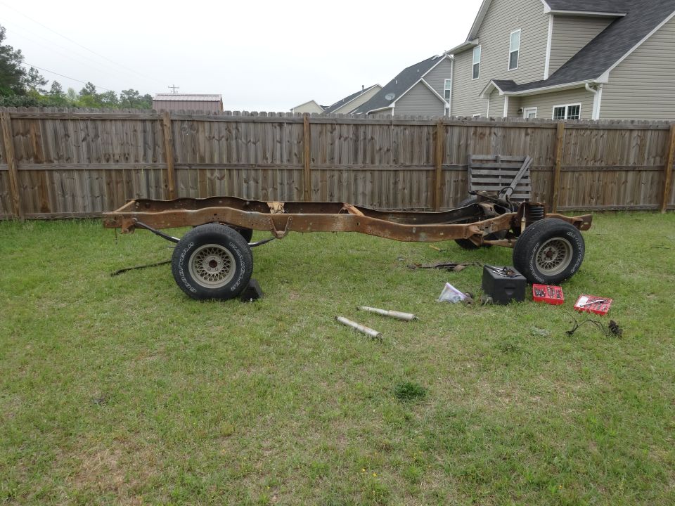 Rusty disassembled 1981 Ford F-100 truck frame with wheels on grass.
