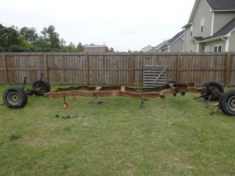 1981 Ford F-100 - A rusty, disassembled truck frame with front and rear axles and wheels attached is displayed on a grassy lawn in front of a wooden fence. T…