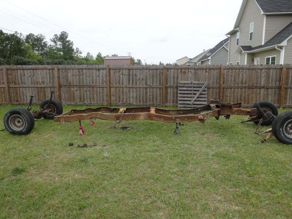 Rusty disassembled 1981 Ford F-100 truck frame with axles and wheels on grass.