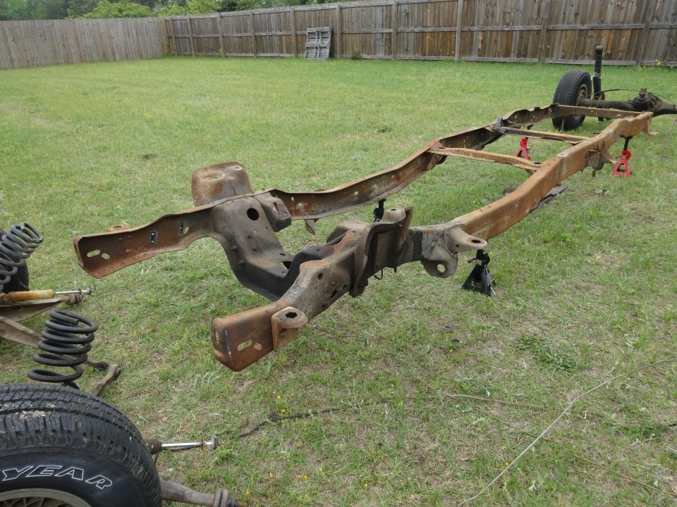 Rusted 1981 Ford F-100 chassis on grass with jack stands and a tire.