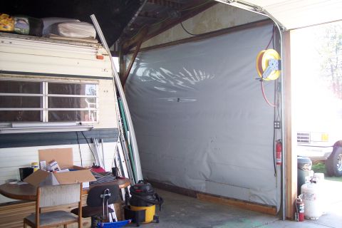 1990 Ford Bronco - A large, grey tarp covers the opening of a garage or workshop, with a vintage RV parked to the left. A yellow hose reel and a red fire exti…