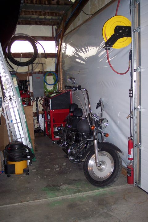 1990 Ford Bronco - A black cruiser-style motorcycle is parked in a garage next to a red sandblaster cabinet and a ladder. A yellow air hose reel is mounted on…