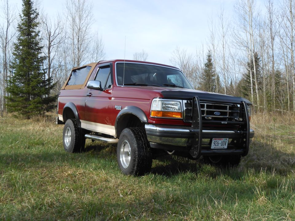 Maroon 1992 Ford Bronco with tan soft top and black brush guard parked in a grassy field.