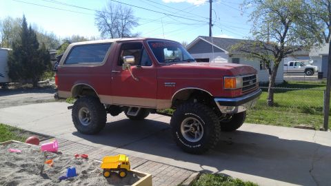 1991 Ford Bronco - A lifted red 1991 Ford Bronco with a tan lower body trim and a tan removable top is parked in a driveway. A sandbox with toys is in the for…