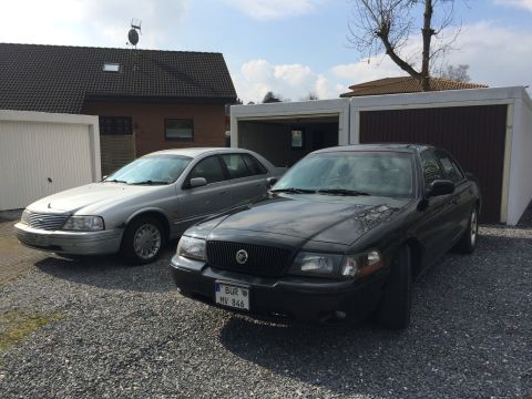 1999 Ford Fairlane - Two sedans are parked in front of garages on a gravel lot. The car in the foreground is black and has a license plate that reads "BUR MV 84…