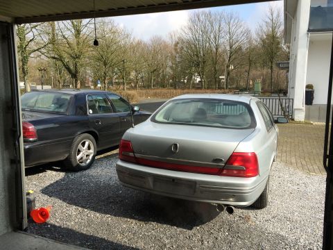 1999 Ford Fairlane - The rear of a silver Ford sedan is parked next to a black sedan in a gravel driveway, viewed from inside a garage.