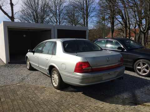 1999 Ford Fairlane - A silver 1999 Ford Fairlane sedan is parked next to a garage. A black sedan is parked to the right of the silver car.