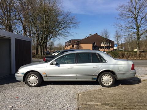 1999 Ford Fairlane - The side view of a silver 1999 Ford Fairlane sedan is parked on a gravel lot next to a garage. The car has a blue decal on the front fender…