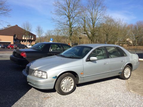 1999 Ford Fairlane - A silver four-door sedan is parked on a gravel lot in front of a dark sedan. The silver sedan is angled towards the viewer, showing its fro…