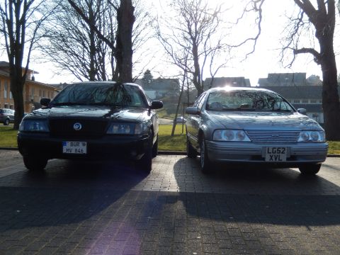 1999 Ford Fairlane - Two sedans are parked side-by-side on a paved surface in front of trees and buildings. The car on the left is black with a license plate re…