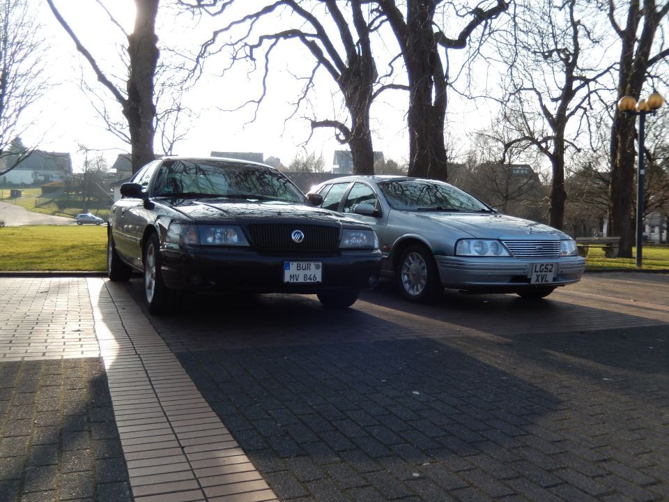 Black and silver sedans parked outdoors with trees and sunlight.