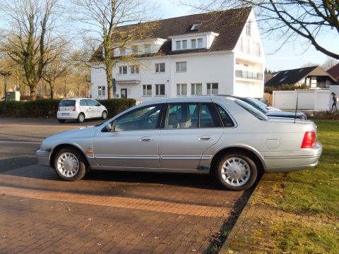 1999 Ford Fairlane - A silver 1999 Ford Fairlane sedan is parked on a brick and grass area in front of a white building. The car is viewed from the side at a 3/…