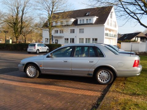 1999 Ford Fairlane - A silver four-door sedan is parked on a brick paver surface next to a grassy area. A white hatchback is visible in the background.