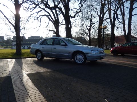 1999 Ford Fairlane - A silver sedan is parked on a brick-patterned driveway with a red sedan visible in the background. Tall trees with bare branches cast shado…