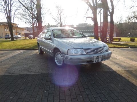 1999 Ford Fairlane - A silver 1999 Ford Fairlane sedan is parked on a brick driveway in front of a building and trees. The car is viewed from a low angle, showi…