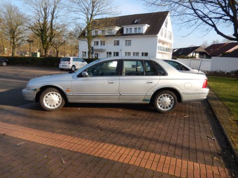 1999 Ford Fairlane - A silver 1999 Ford Fairlane sedan is parked on a brick paver surface in front of a white building. The car is viewed from the side, showing…