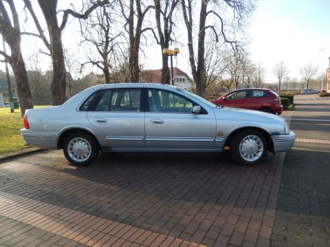 1999 Ford Fairlane - The side profile of a silver 1999 Ford Fairlane sedan is parked on a brick-paved lot. A red hatchback is parked behind it, and bare trees a…
