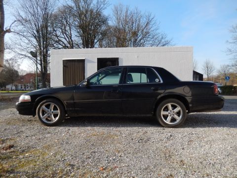 1999 Ford Fairlane - The side view of a black four-door sedan parked on a gravel lot in front of a white building. The car has silver multi-spoke wheels and tin…