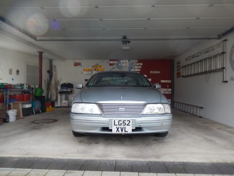 1999 Ford Fairlane - A silver sedan is parked head-on in a garage, facing the camera. The vehicle has a chrome grille and a license plate reading "LG52 XVL".