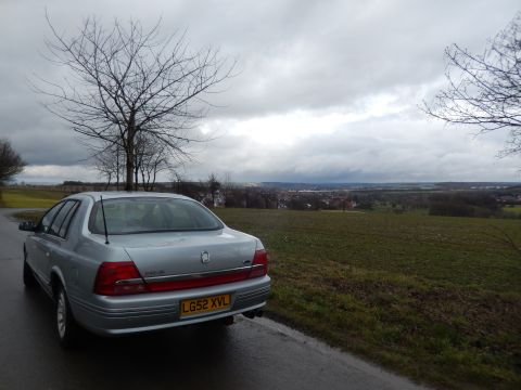 1999 Ford Fairlane - A silver sedan is parked on the side of a road with a scenic view of a town in the distance under a cloudy sky.
