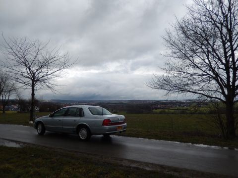 1999 Ford Fairlane - A silver 1999 Ford Fairlane sedan is captured from the rear as it drives along a wet road, with a scenic, overcast landscape in the backgro…