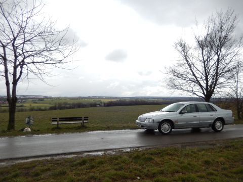 1999 Ford Fairlane - A silver sedan is parked on the side of a road with a scenic view of fields and trees under a cloudy sky.