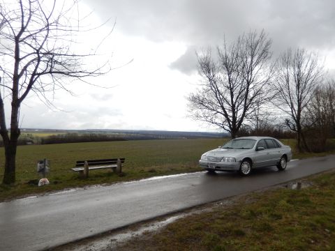 1999 Ford Fairlane - A silver sedan drives on a wet road with a field and bare trees under a cloudy sky.