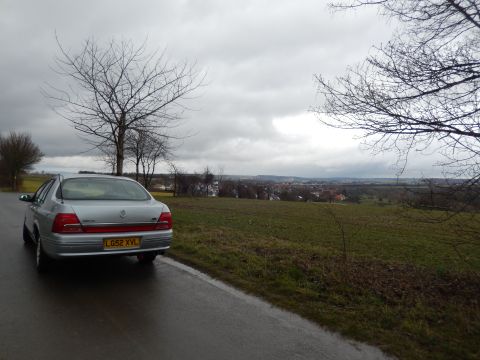 1999 Ford Fairlane - A silver sedan is parked on the side of a wet road, with a town visible in the distance under a cloudy sky.