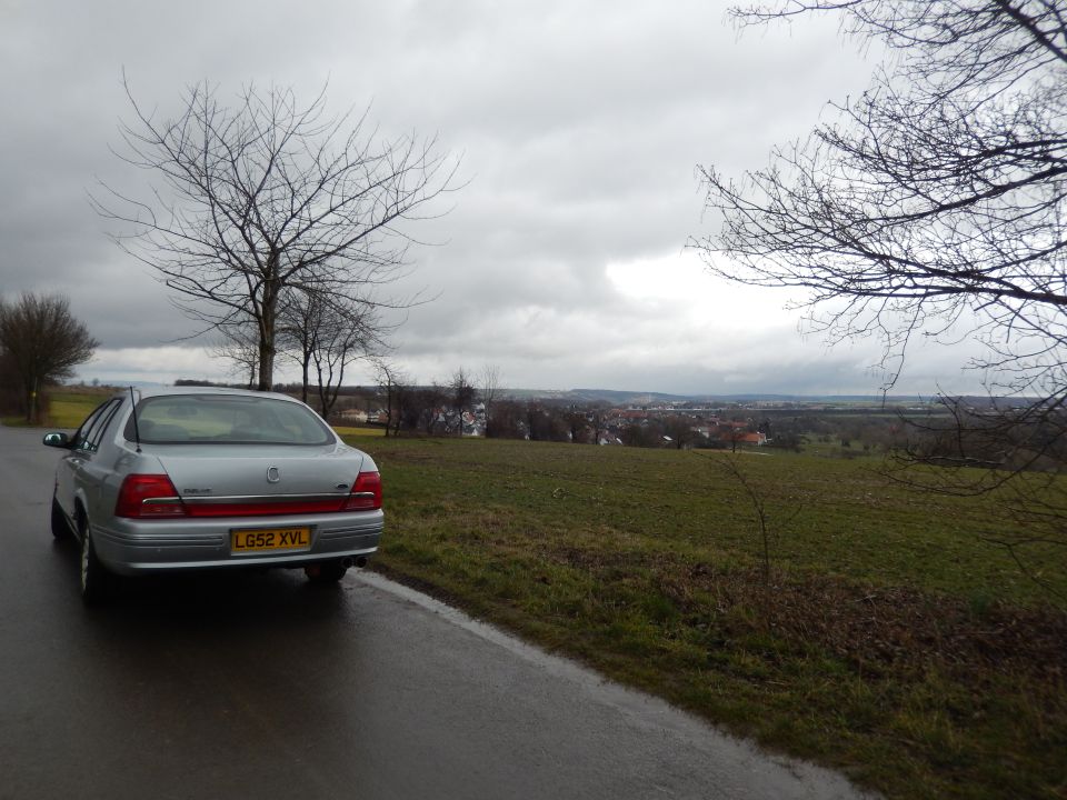Silver sedan parked on the side of a wet road with a town in the distance under a cloudy sky