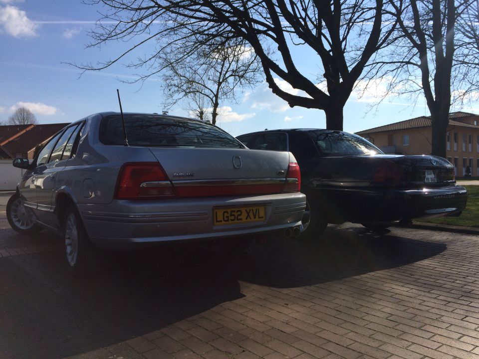 Silver sedan and black sedan parked side-by-side on a brick surface with trees overhead