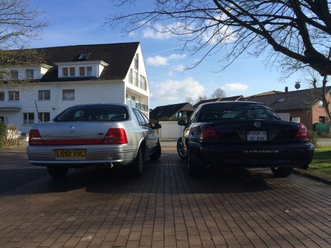1999 Ford Fairlane - Two sedans, a black Mercury Marauder and a silver sedan, are parked on a brick driveway in front of houses.