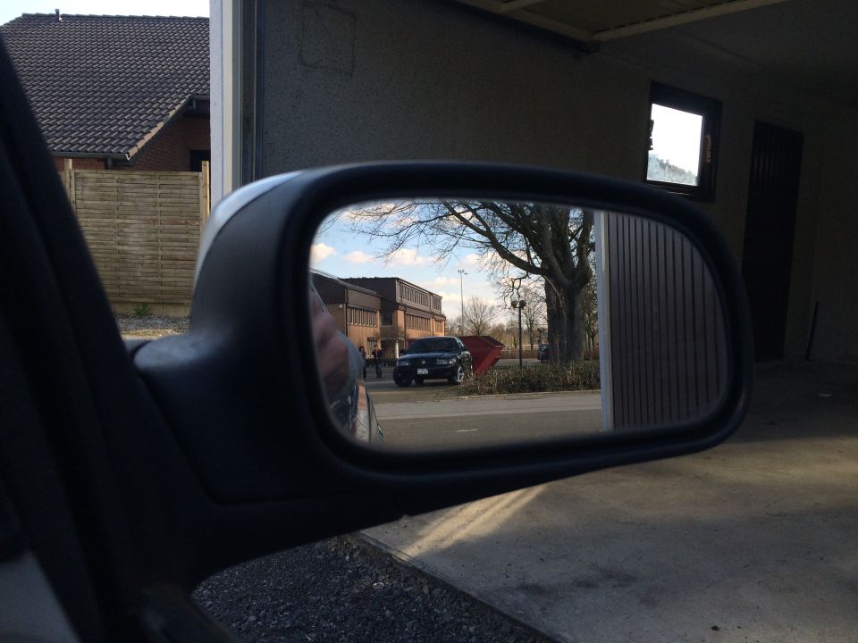 Side mirror reflection shows a dark SUV parked on a street with a building and trees in the background