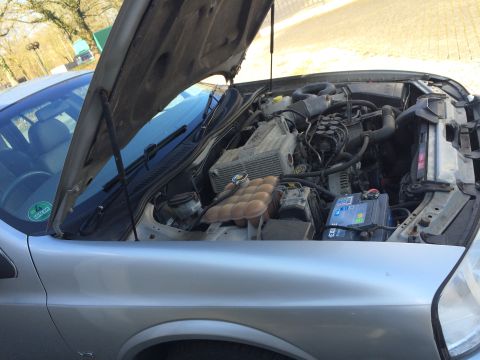 1999 Ford Fairlane - Engine bay of a silver car, showing the battery, coolant reservoir, and engine components with the hood open.