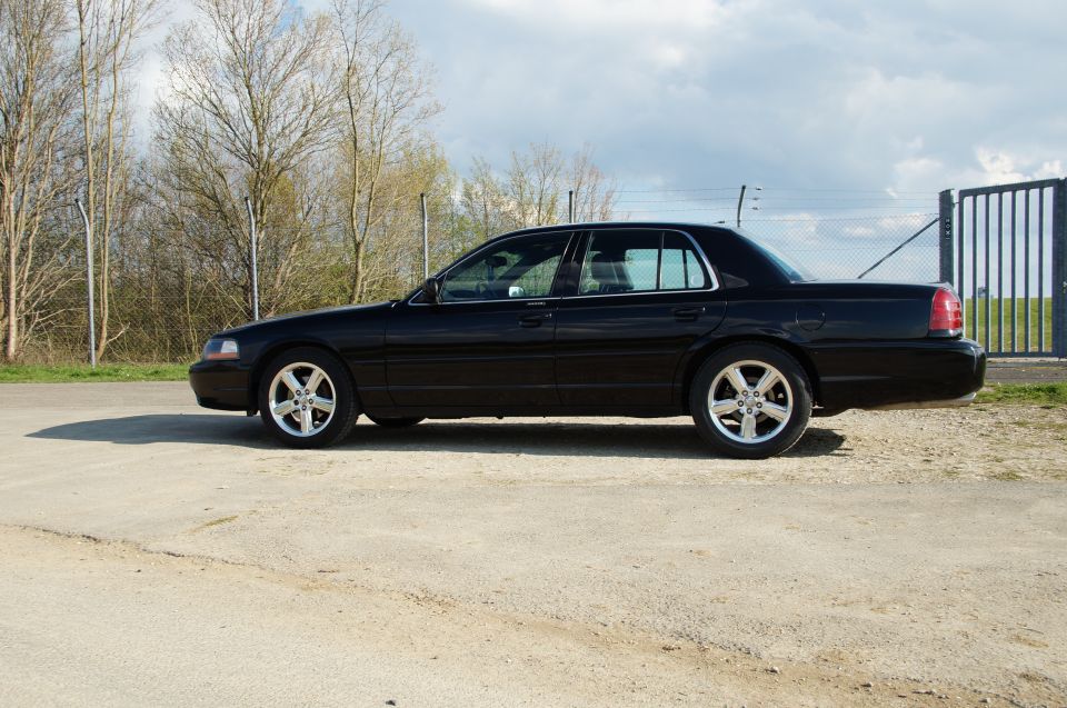 Black sedan parked on dirt lot, side view. It has aftermarket wheels