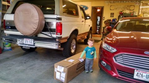 1994 Ford Bronco - A child stands between a beige Ford Bronco and a red Ford sedan in a garage.